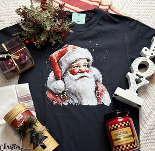 Black t-shirt with Santa Claus graphic, Christmas-themed candles, and decorative items on a textured surface.